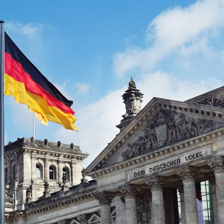 Bundesflagge vor dem Reichstagsgebäude in Berlin unter blauem Himmel.